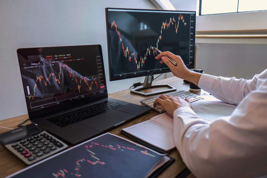 Trader analyzing forex and stock charts on dual monitors and a laptop, using candlestick patterns and technical indicators at a desk with notebook and calculator.