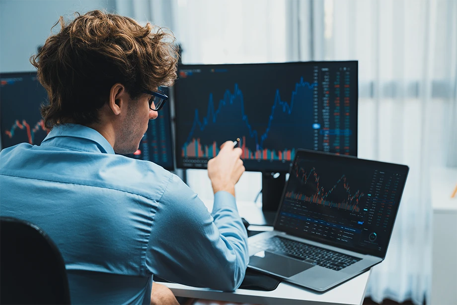 Trader analyzing multiple financial charts on laptop and monitors, reviewing market trends for trading decisions.